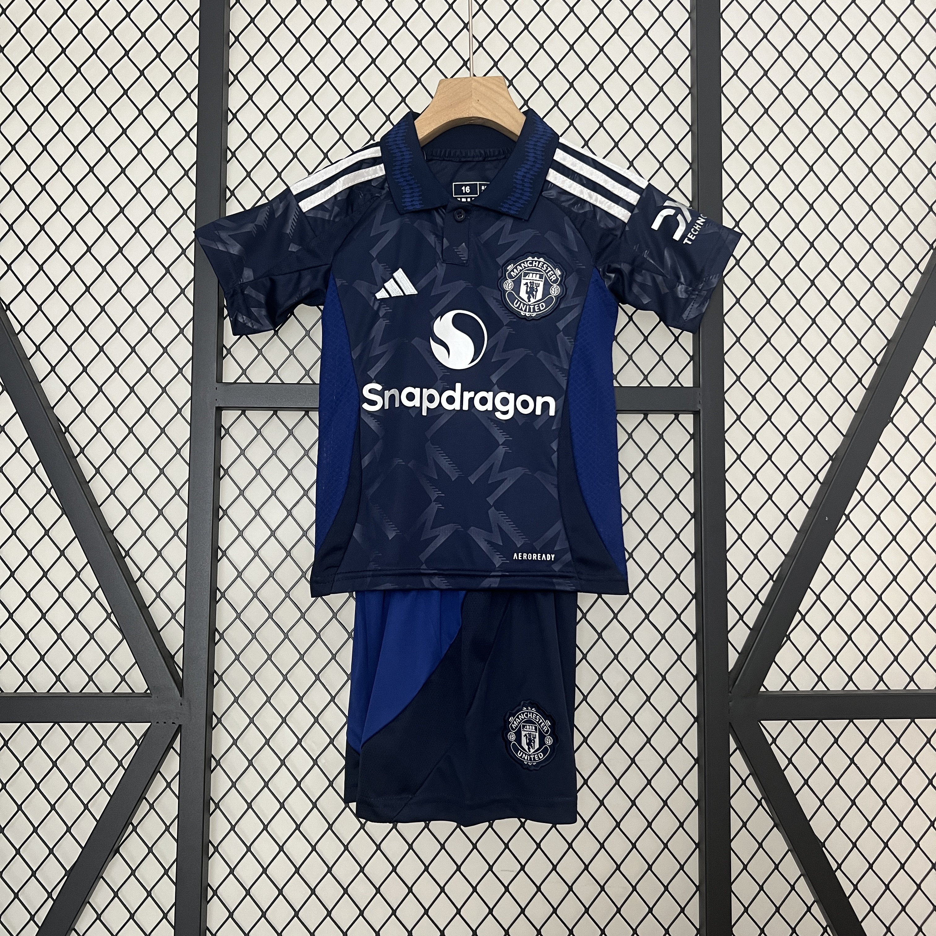 Kids Manchester United Away Kit 2024/25 Kids Manchester United Away Kit 2024/25 on hanger showing navy monogram shirt with royal blue polo collar and shorts