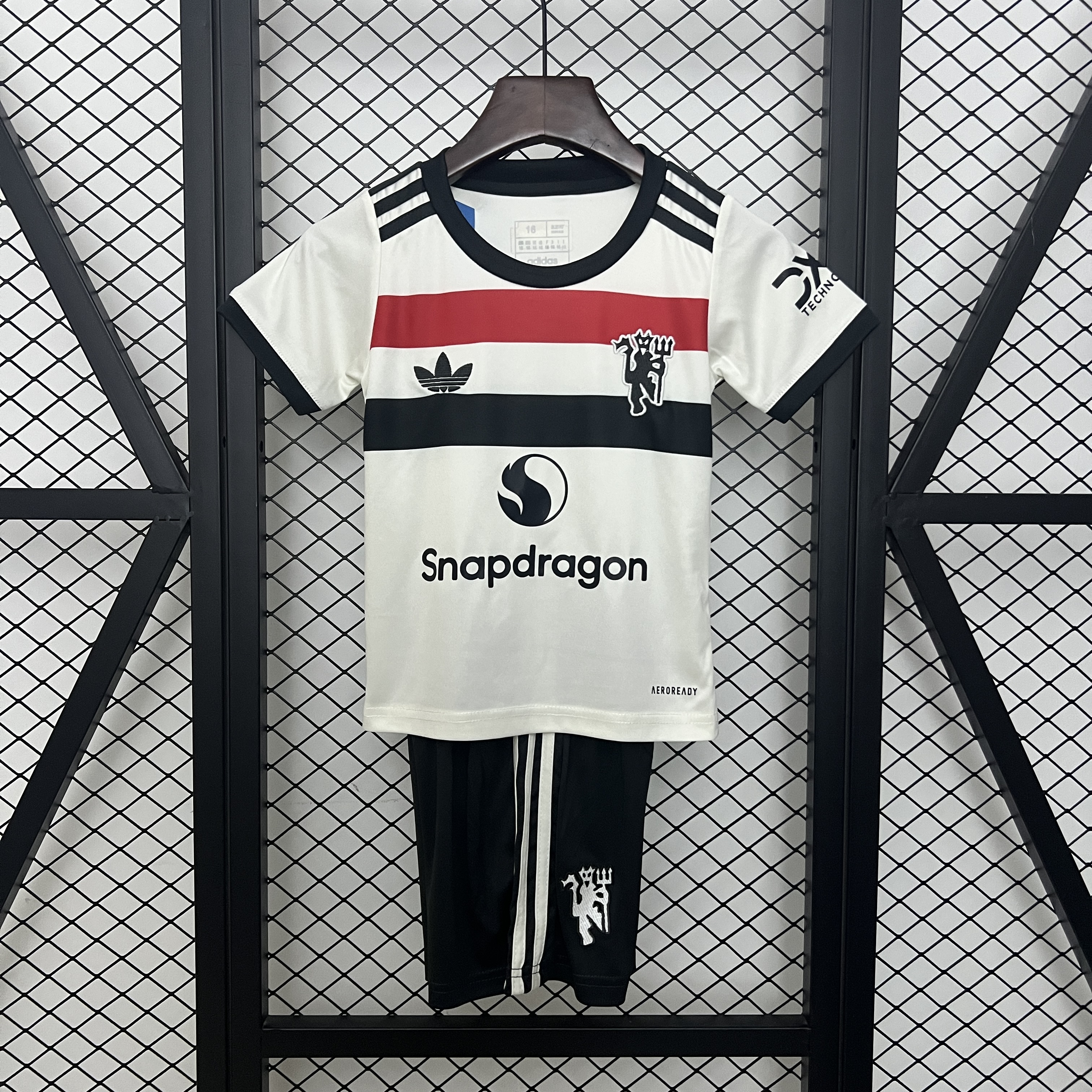 Kids Manchester United Third Kit 2024/25 Kids Manchester United Third Kit 2024/25 on hanger showing off-white jersey with tri-color chest band and black shorts